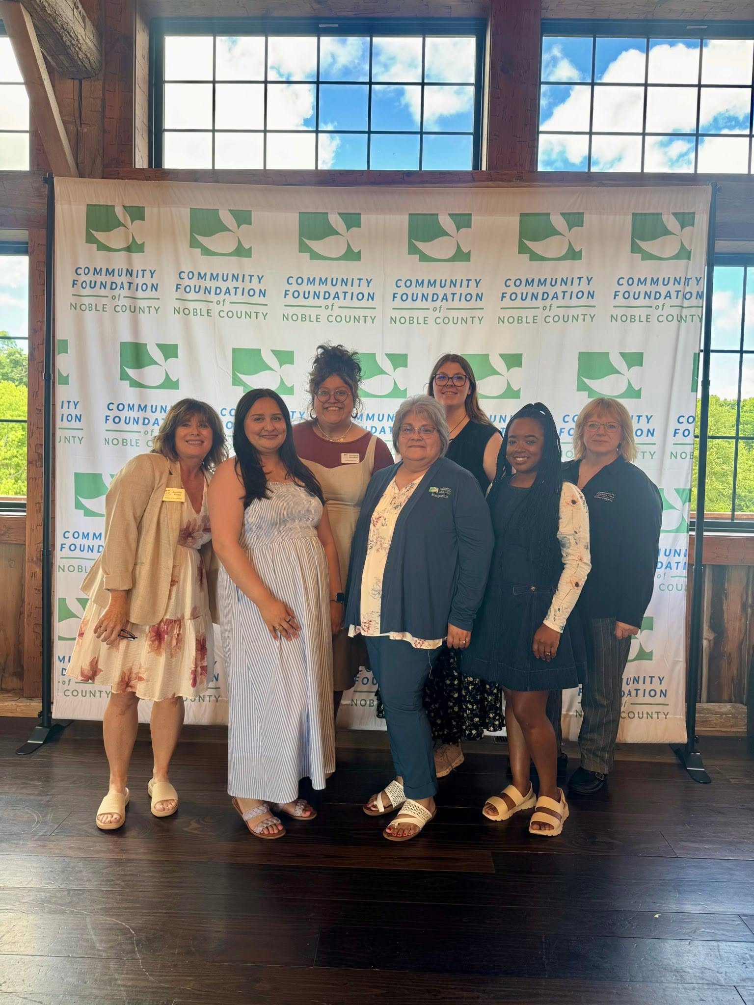 Seven women stand together smiling in front of a Community Foundation of Noble County backdrop, inside a wooden building with large windows showing a blue sky and clouds.