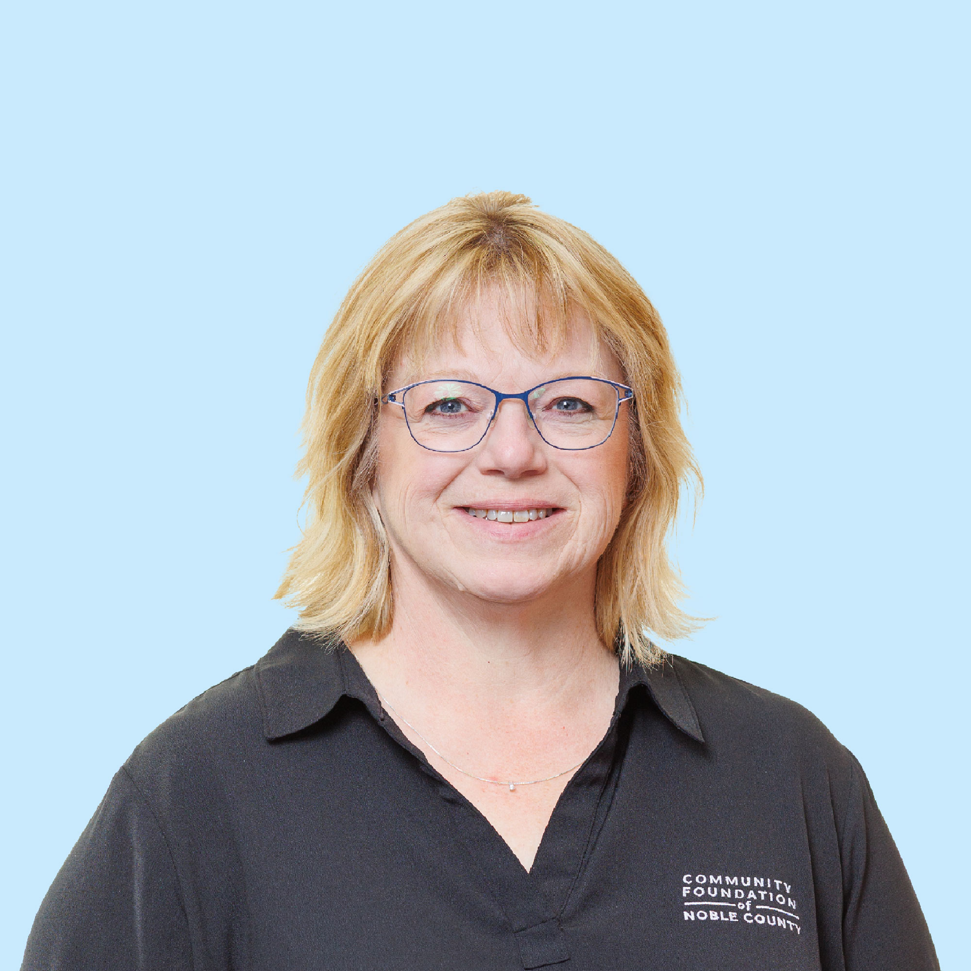 Smiling woman with blonde hair, glasses, and a black shirt that says "Community Foundation of Noble County," standing against a light blue background.