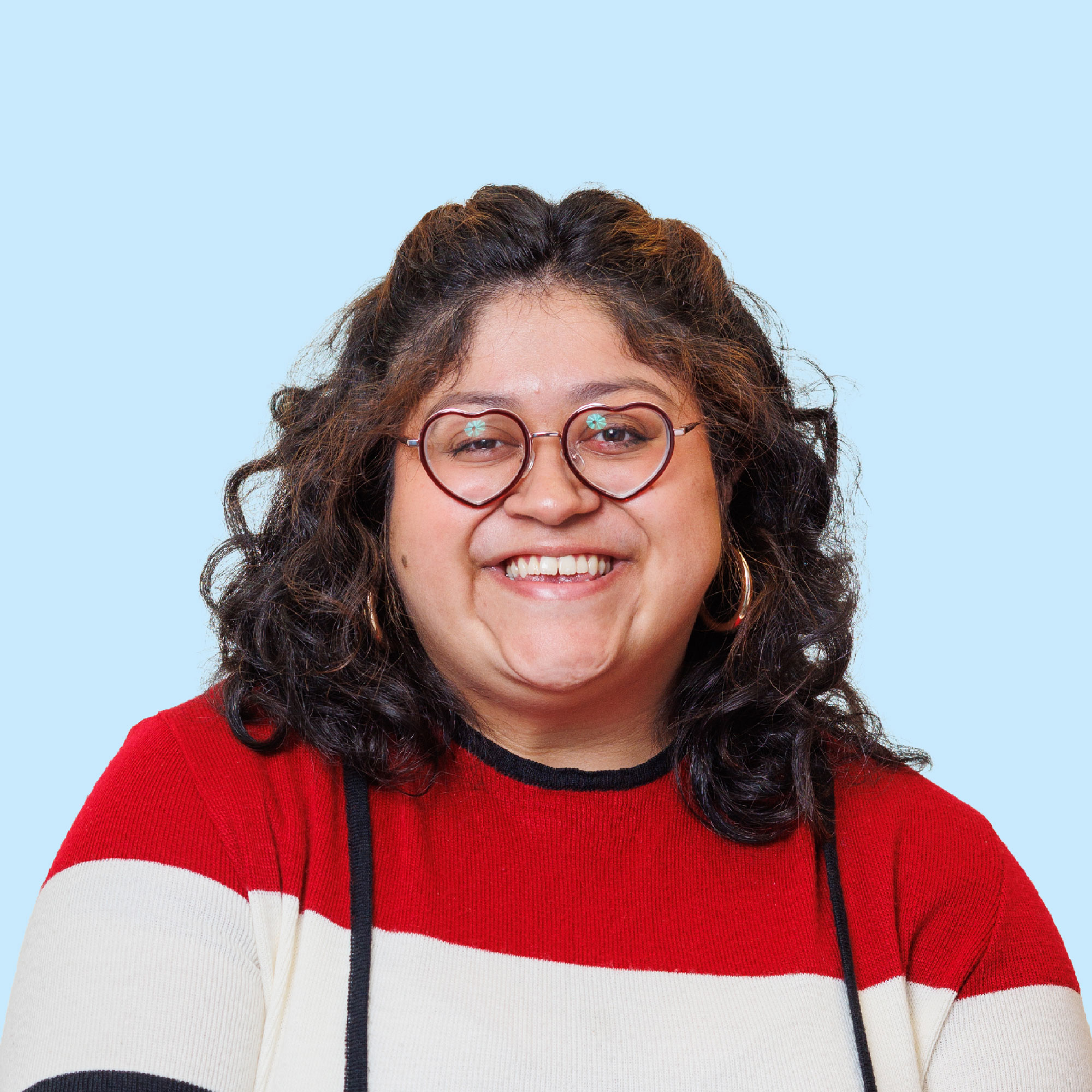 Smiling person with curly dark hair and glasses, wearing a red, white, and black sweater, posed against a light blue background.