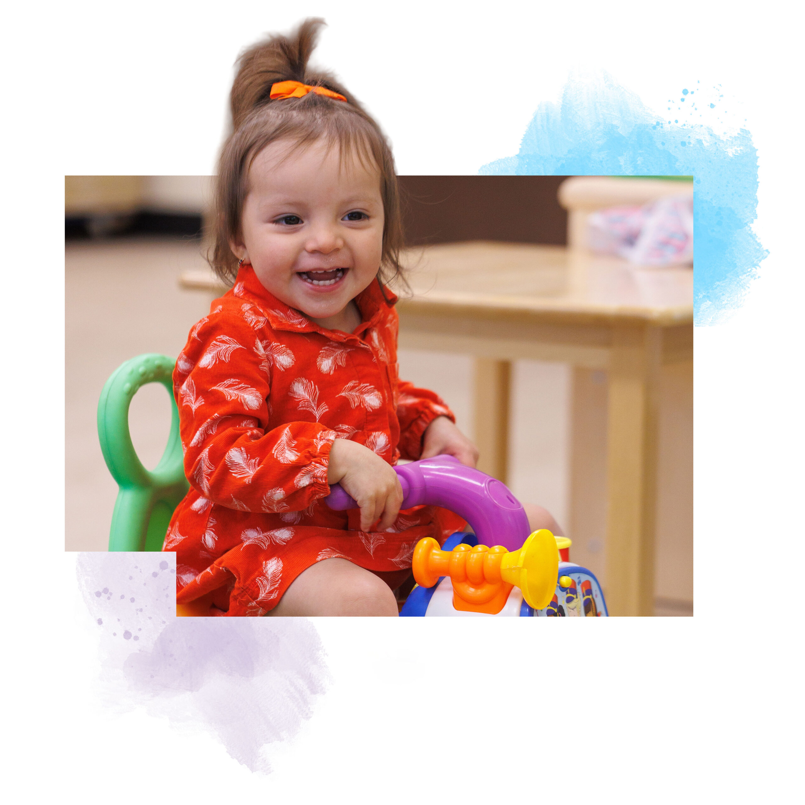 Smiling toddler in a red patterned dress sits on a colorful toy bike indoors, with a table and pastel brushstroke design in the background.